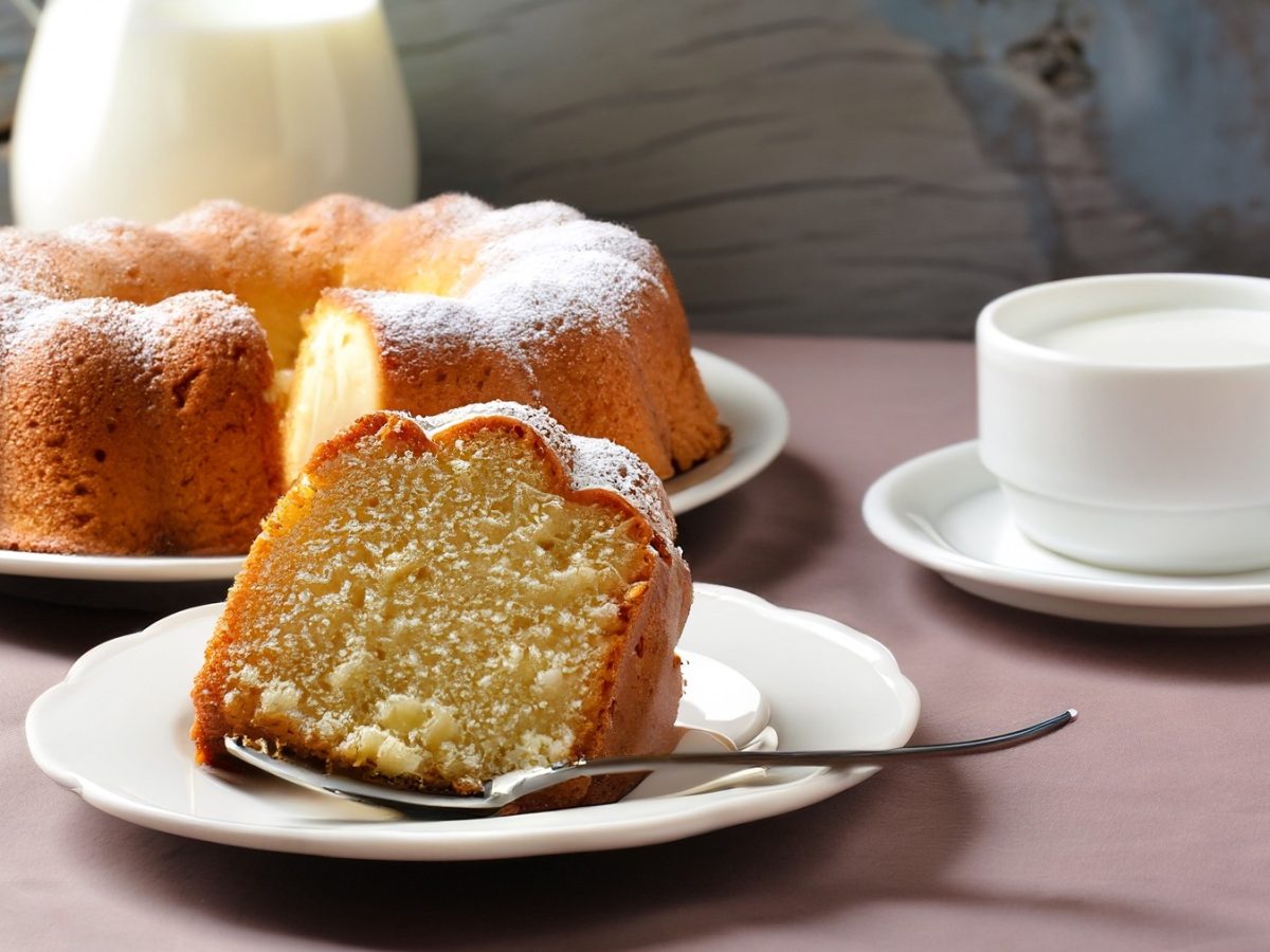 Ein frisch gebackener französischer Joghurtkuchen mit Puderzucker, ein Stück davon auf einem Teller mit Löffel, daneben eine Tasse Milch.