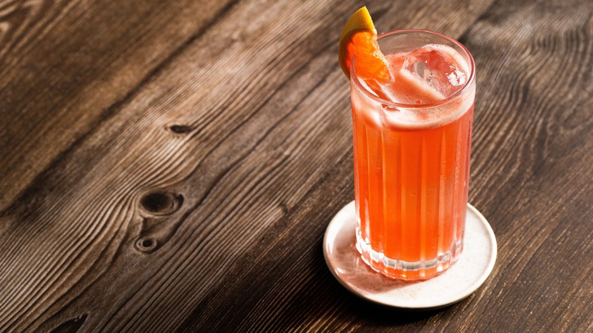 Ein leuchtender Garibaldi-Cocktail mit Eiswürfeln und Orangengarnitur steht auf einer rustikalen Holzplatte in warmem Licht.