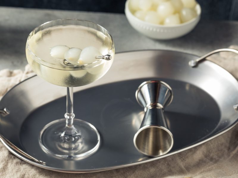 Ein klarer Gibson mit drei Perlzwiebeln auf einem Cocktailspieß, serviert auf einem silbernen Tablett mit Jigger und Schale im Hintergrund.