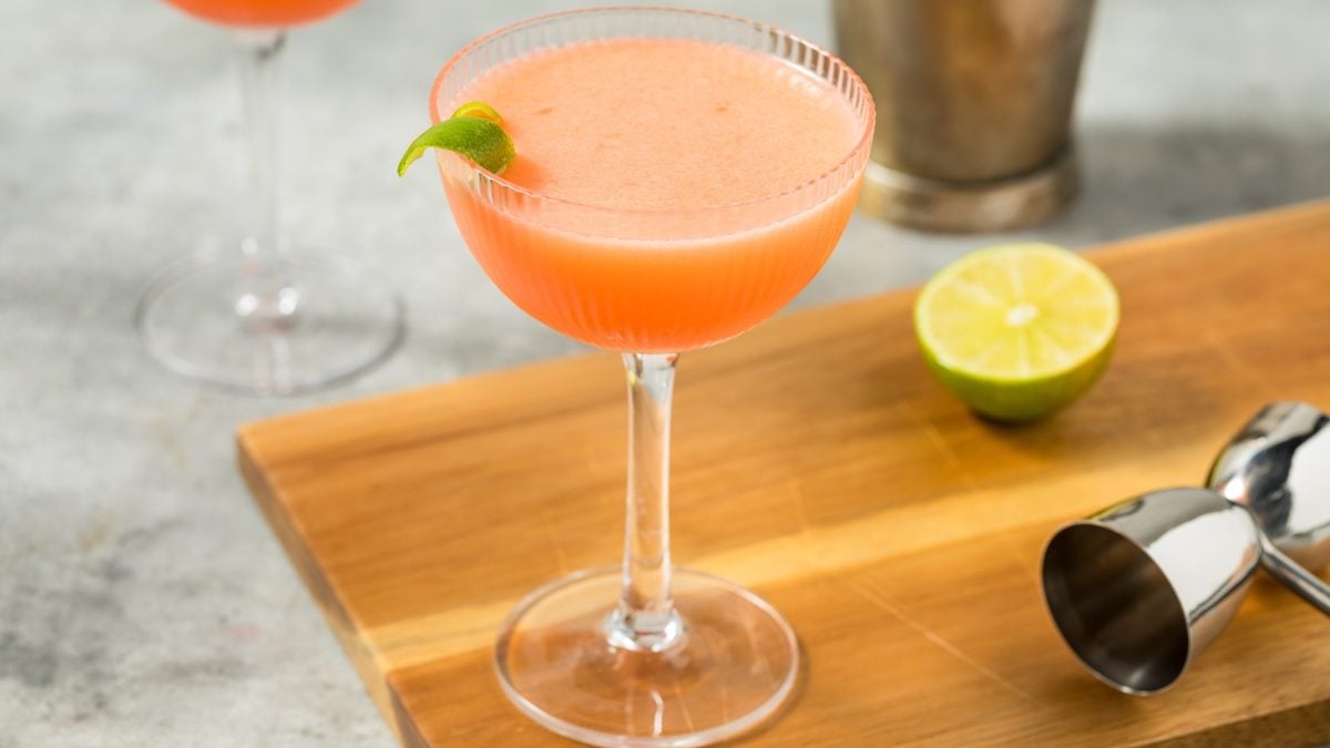 Frischer Great Silence mit schaumiger Oberfläche in edlem Glas, dekoriert mit Limettenzeste, auf Holzbrett mit Limette und Barzubehör.