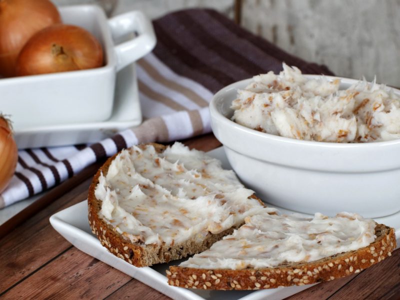 Frontal: Auf einem Holztisch stehen eine helle, runde Schüssel mit Griebenschmalz. Daneben liegen zwei halbe Brotscheiben mit dem Aufstrich. Im Hintergrund sind frische Zwiebeln.