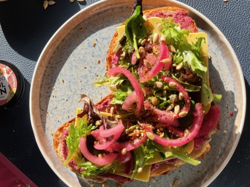 Bunt belegtes Brot mit Rote-Bete-Aufstrich, Käse, frischem Salat, eingelegten Zwiebeln und Sonnenblumenkernen auf Keramikteller.