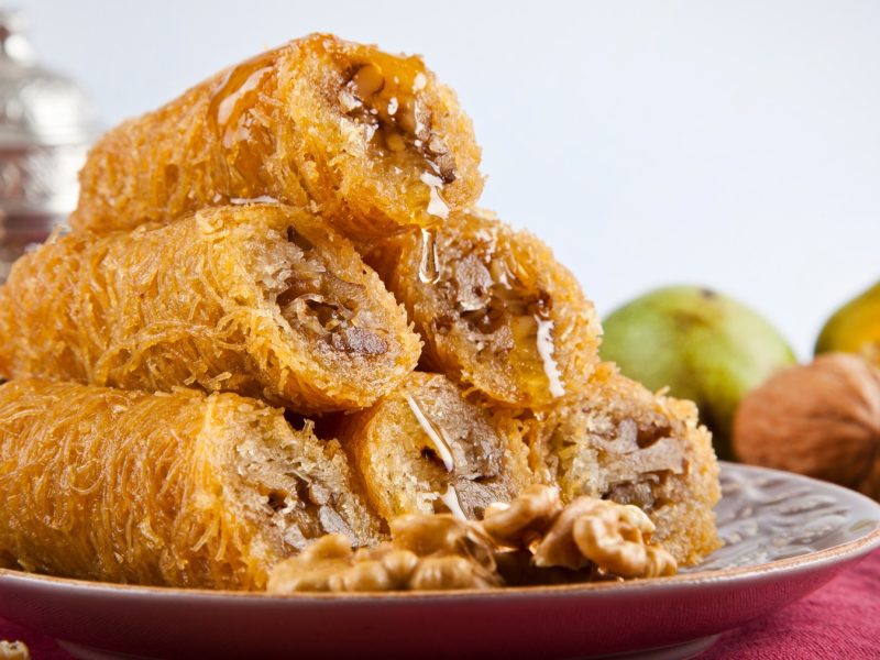 Knusprige, goldbraune Kadayıf Dolması mit Walnussfüllung, überzogen mit Honig, auf einem Teller mit roten Tuch und Nüssen im Hintergrund.