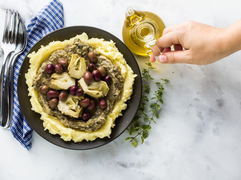 Hand gießt Öl über Kartoffelpüree mit Artischocken, Oliven und Baba Ganoush auf schwarzem Teller, daneben blaue Serviette.