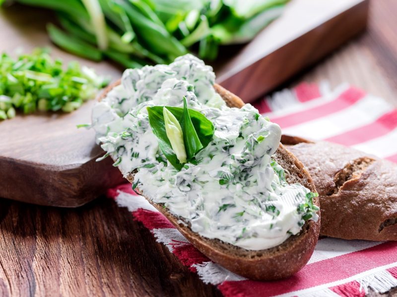 Draufsicht: Auf einem Holztisch liegen ein Brot mit Kräuterquark und ein Küchenhandtuch. Drum herum sind frische Kräuter.