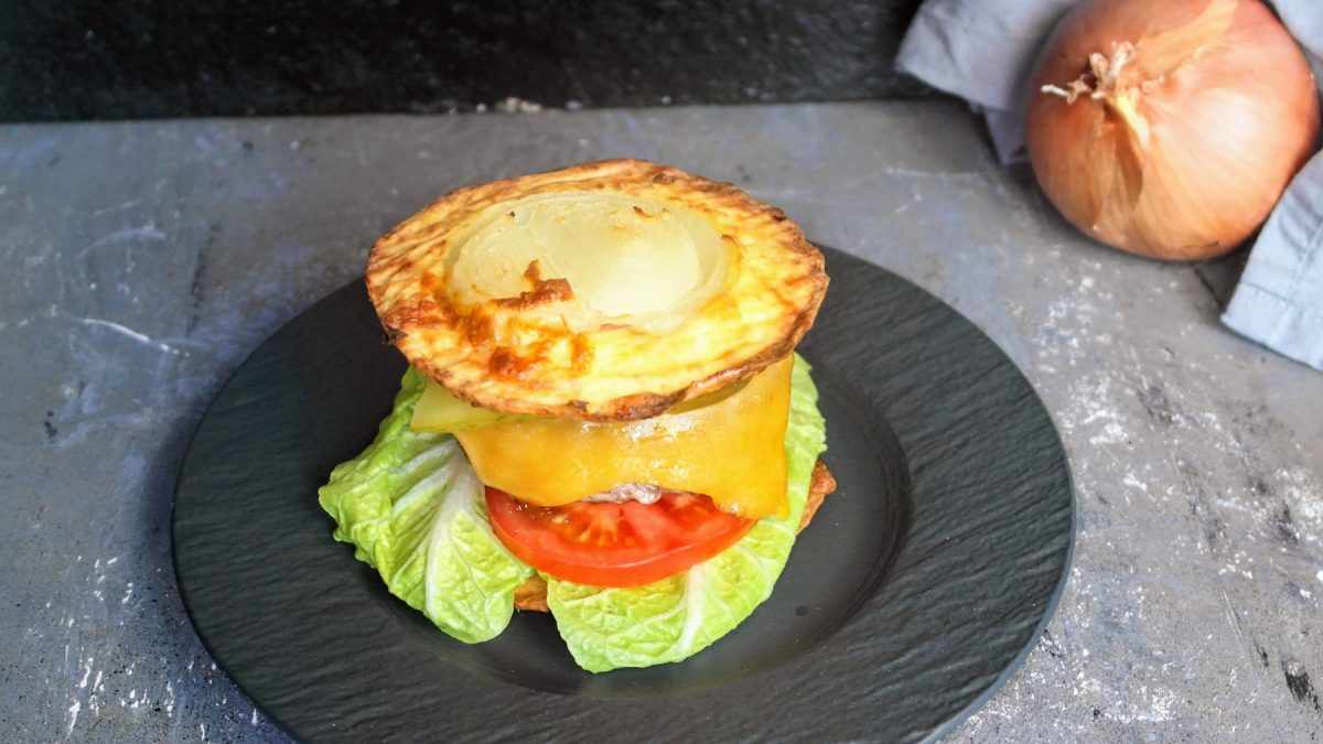 Der Low-Carb-Burger auf einem schwarzen Teller von schräg oben fotografiert. Alles auf einem Steintisch mit einem grauen Tuch und einer Zwiebel im Hintergrund.