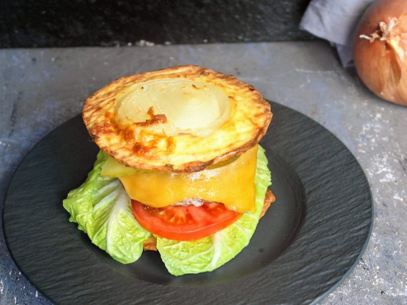 Der Low-Carb-Burger auf einem schwarzen Teller von schräg oben fotografiert. Alles auf einem Steintisch mit einem grauen Tuch und einer Zwiebel im Hintergrund.