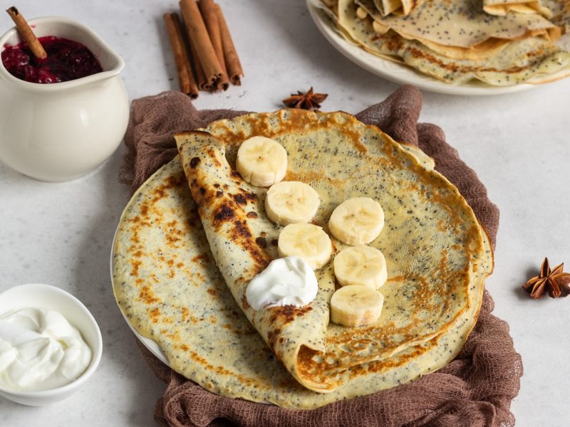 Nahaufnahme eines Mohnpfannkuchens mit karamellisierten Stellen, serviert mit Bananen, Joghurt und Gewürzen auf hellem Steinhintergrund.