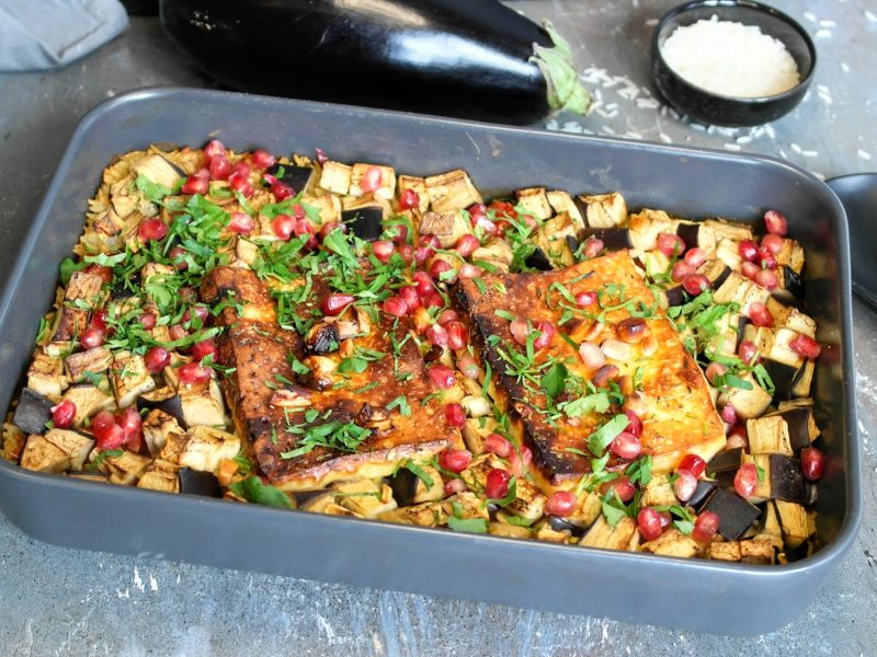 Eine graue Auflaufform mit dem Reisgericht auf einem Steintisch. Daneben ein schwarzer Kochlöffel und dahinter eine Aubergine sowie ein Schälchen mit Reis. Alles von schräg oben fotografiert.