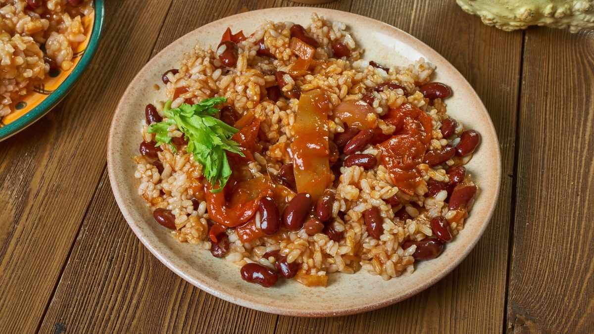 Draufsicht: Eine runder Teller auf einem Untergrund aus Holz. Auf dem Teller ist ein Gericht mit Bohne, Reis, Tomaten und Paprika.