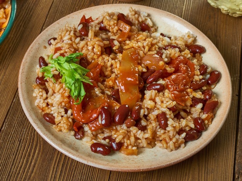 Draufsicht: Eine runder Teller auf einem Untergrund aus Holz. Auf dem Teller ist ein Gericht mit Bohne, Reis, Tomaten und Paprika.