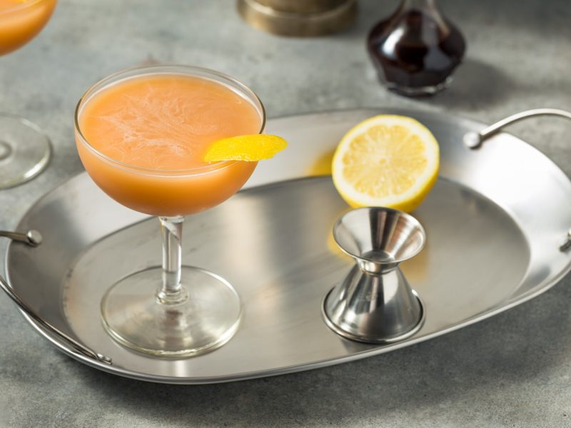 Ein Pantheon-Cocktail mit schaumiger Oberfläche, serviert mit Zitronenzeste, auf einem Tablett mit Jigger und Zitronenhälfte.