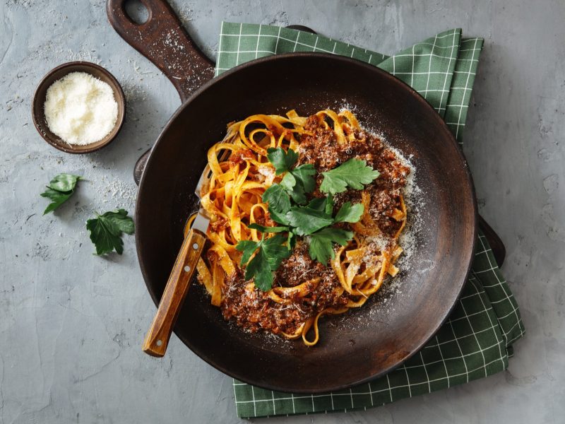 Ein dunkelbrauner Teller mit der Bolognese auf einem hellgrauen Tisch mit Parmesan und Basilikum.