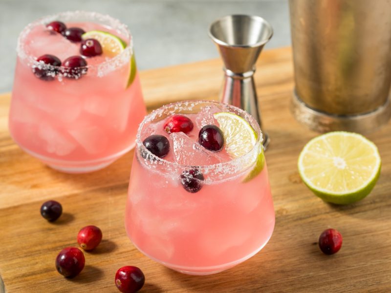 Eisgekühlte Cranberry-Margaritas in Gläsern mit gesalzenem Rand, Cranberrys und Limetten, arrangiert auf einem Holzbrett.