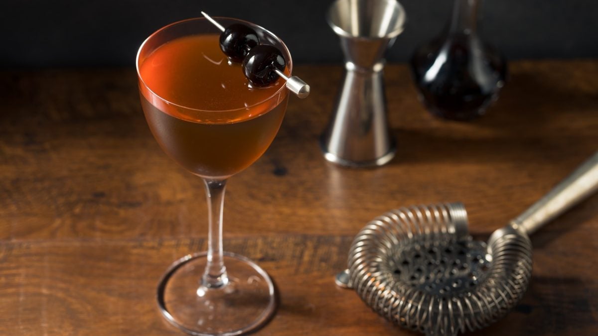 Ein klassischer Rob-Roy-Cocktail in einem eleganten Glas mit zwei dunklen Kirschen auf einem Metallspieß, serviert auf Holz.