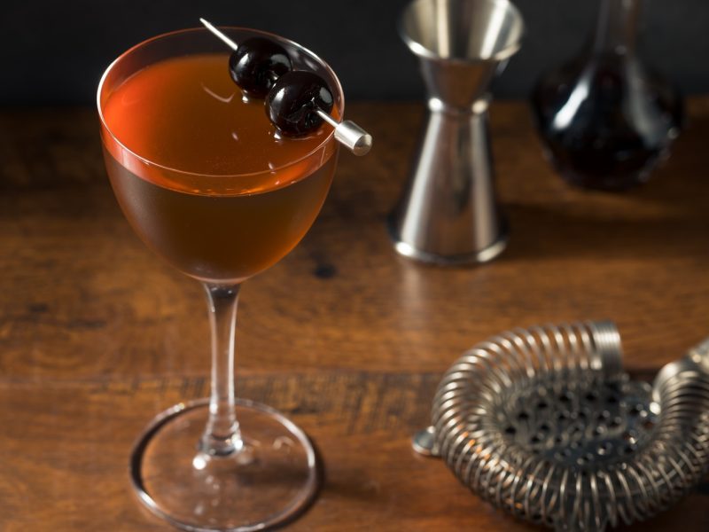 Ein klassischer Rob-Roy-Cocktail in einem eleganten Glas mit zwei dunklen Kirschen auf einem Metallspieß, serviert auf Holz.