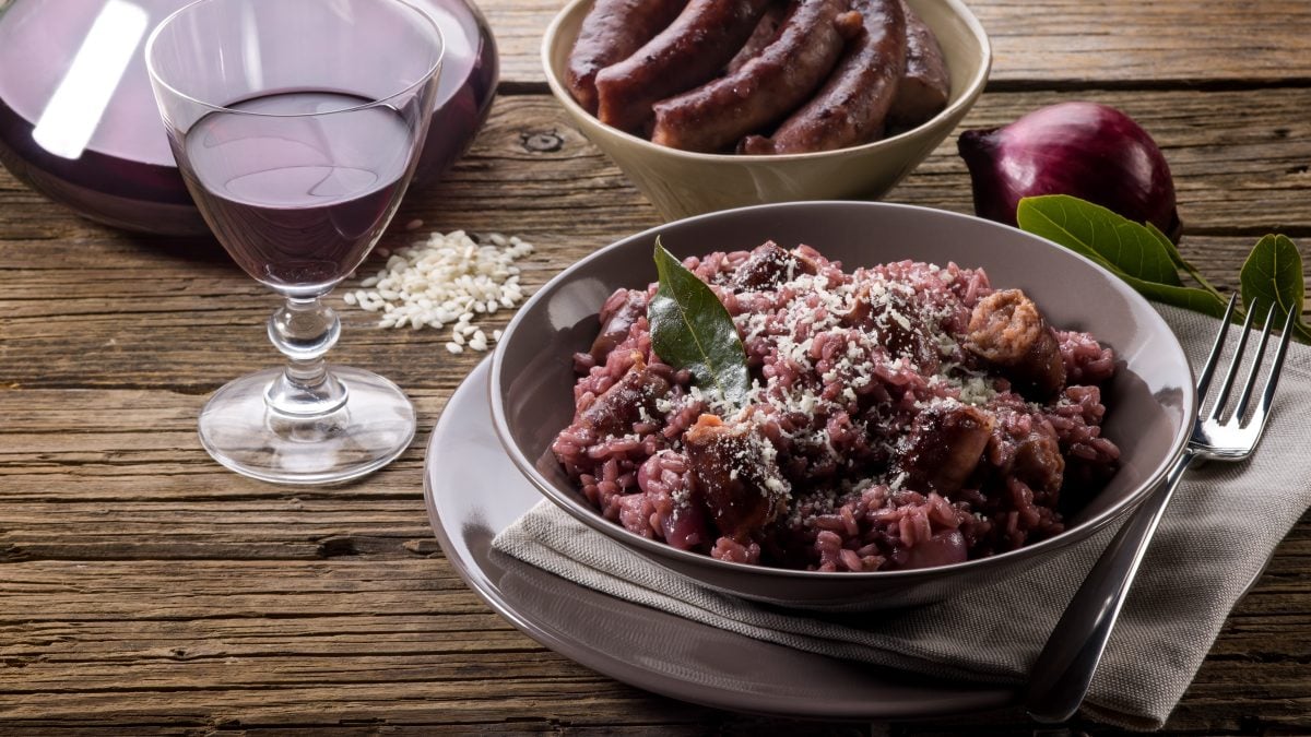 Draufsicht, schräg: Ein runder Teller mit einem Rotwein-Risotto und Wurst. Daneben stehen ein Glas und eine Karaffe mit Rotwein. Im Hintergrund ist eine Schale mit Würsten vom Metzger.