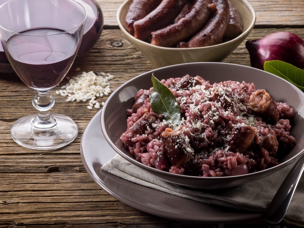 Draufsicht, schräg: Ein runder Teller mit einem Rotwein-Risotto und Wurst. Daneben stehen ein Glas und eine Karaffe mit Rotwein. Im Hintergrund ist eine Schale mit Würsten vom Metzger.