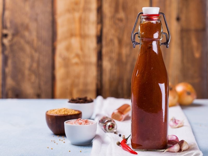 Eine Flasche mit der abgefüllten Schaschlik_Sauce auf einem grauen Tisch und hölzernem Hintergrund. Alles von der Seite fotografiert. Um die Flasche herum liegen noch Kochutensilien und Zutaten.