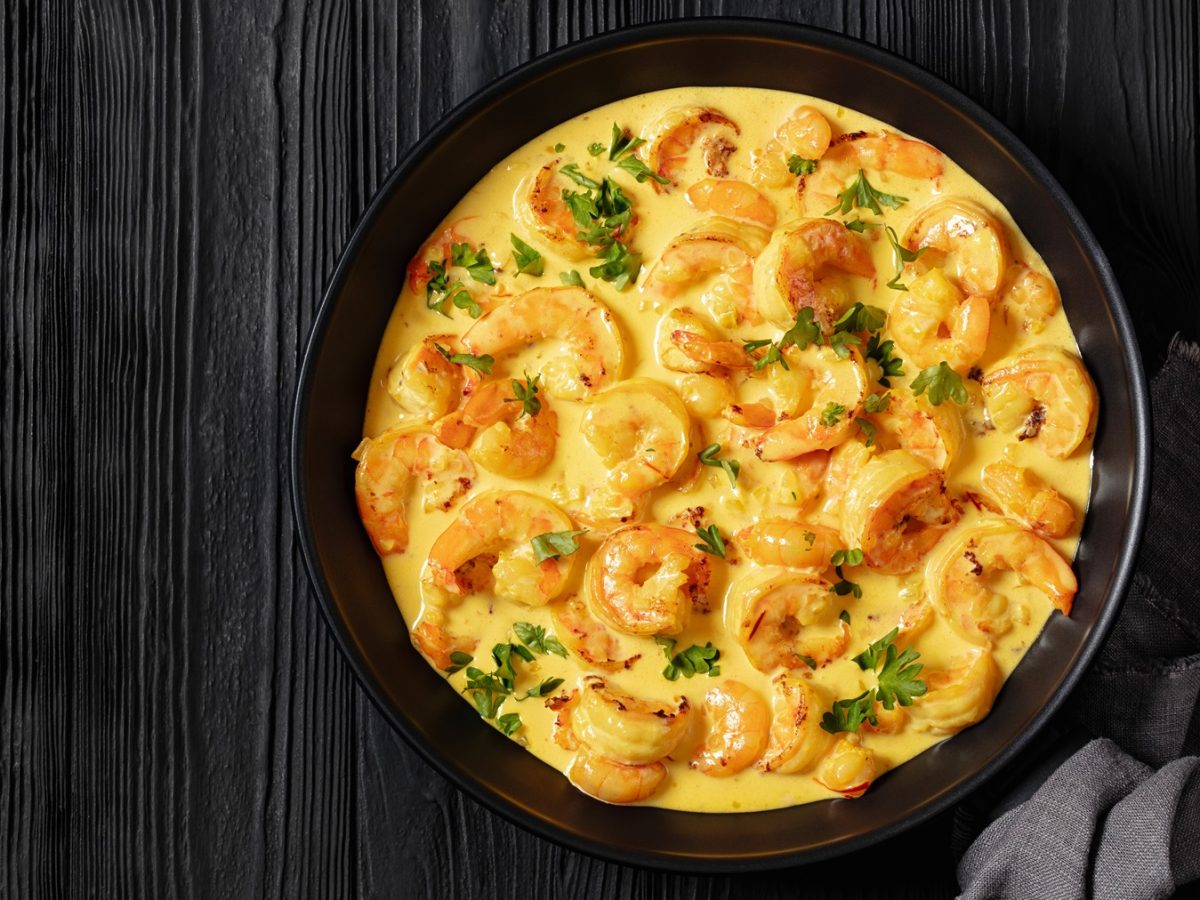 Saftige Shrimps in Safran-Sauce mit Kräutern angerichtet, in einer schwarzen Schüssel auf rustikalem dunklem Holzuntergrund.