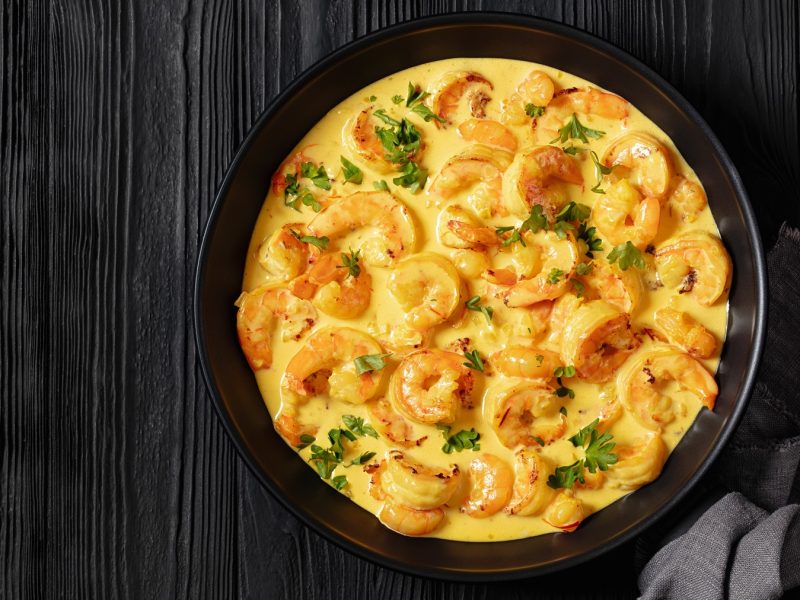 Saftige Shrimps in Safran-Sauce mit Kräutern angerichtet, in einer schwarzen Schüssel auf rustikalem dunklem Holzuntergrund.