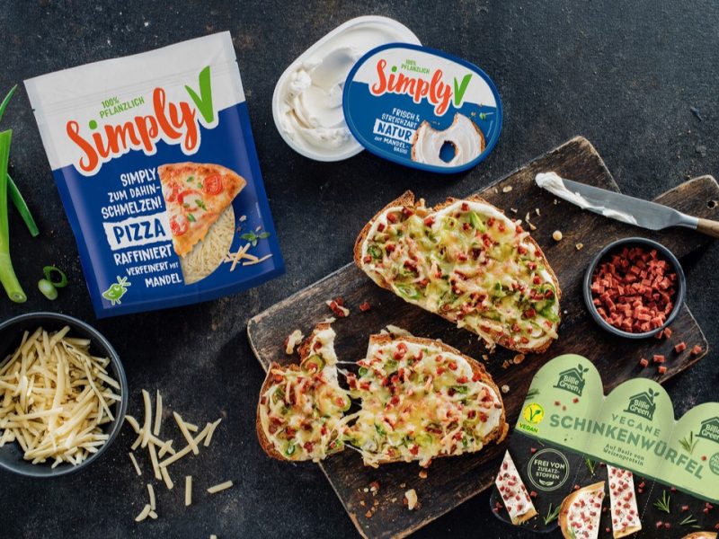 Vegane Flammkuchen-Stulle mit geschmolzenem Käse, Frühlingszwiebeln und Schinkenwürfeln neben Simply V und Billie Green Produkten.