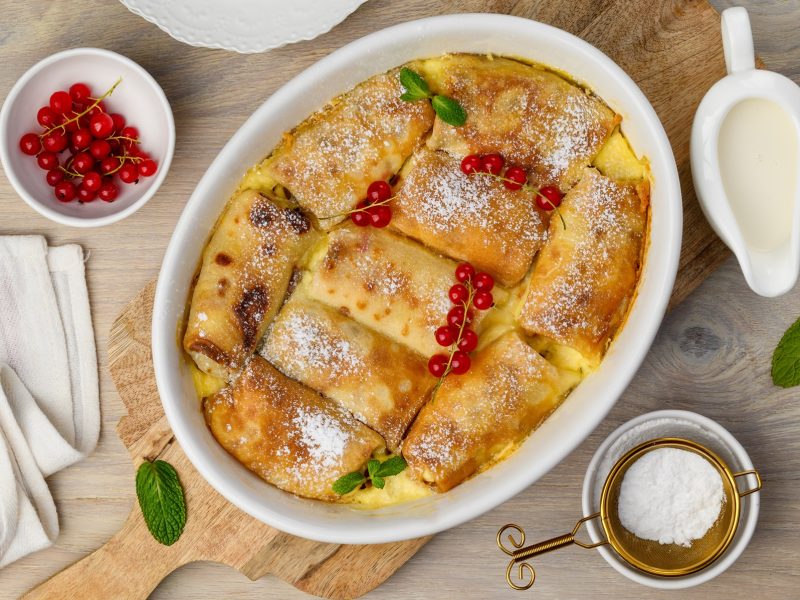 Frisch gebackene goldbraune Topfenpalatschinken in einer weißen Auflaufform, mit Puderzucker bestäubt und mit roten Johannisbeeren garniert.