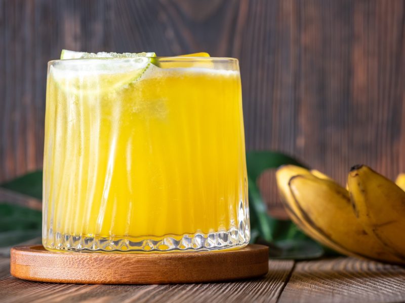Ein erfrischender, goldgelber Yellow-Bird-Cocktail mit Limettenscheiben in einem Kristallglas auf Holzunterlage, im Hintergrund reife Bananen und grüne Blätter.