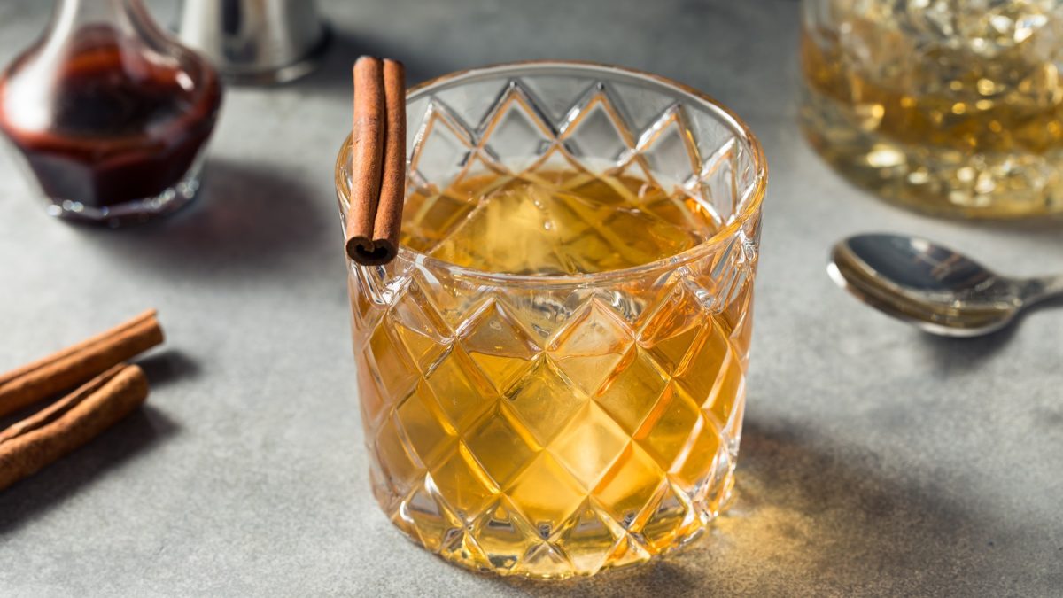 Kristallglas mit Old Fashioned, Eiswürfeln und Zimtstange auf grauem Untergrund, daneben Zimtstangen und ein Löffel.