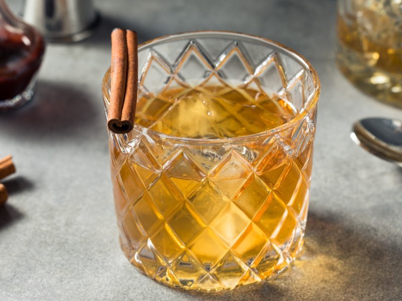 Kristallglas mit Old Fashioned, Eiswürfeln und Zimtstange auf grauem Untergrund, daneben Zimtstangen und ein Löffel.