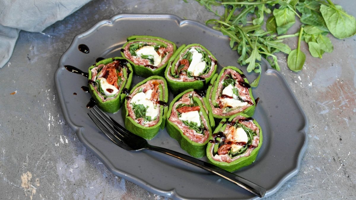 Ein grauer Teller mit der geschnittenen Antipasti-Rolle auf einem Steintisch von schräg oben fotografiert. Im Hintergrund unscharf Rucola, Babyspinat sowie ein graues Tuch erkennbar.