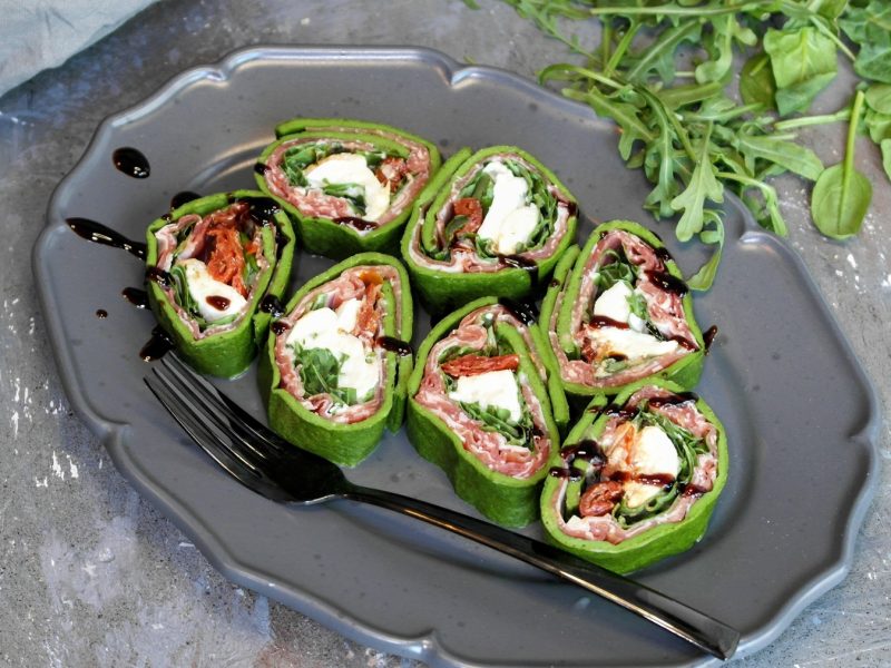 Ein grauer Teller mit der geschnittenen Antipasti-Rolle auf einem Steintisch von schräg oben fotografiert. Im Hintergrund unscharf Rucola, Babyspinat sowie ein graues Tuch erkennbar.