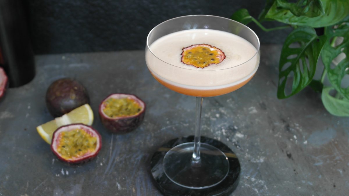 Der Drink auf einem grauen Steintisch mit Passionsfrucht als Deko und schwarzem Hintergrund.