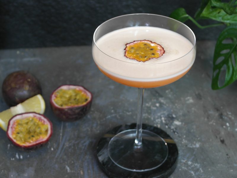 Der Drink auf einem grauen Steintisch mit Passionsfrucht als Deko und schwarzem Hintergrund.