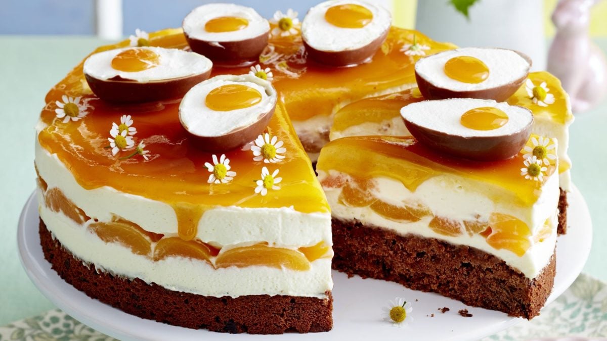 Saftige Aprikosen-Schmand-Torte mit Schokoboden, Sahnecreme und Aprikosenguss, dekoriert mit Schoko-Ostereiern und weißen Gänseblümchen.