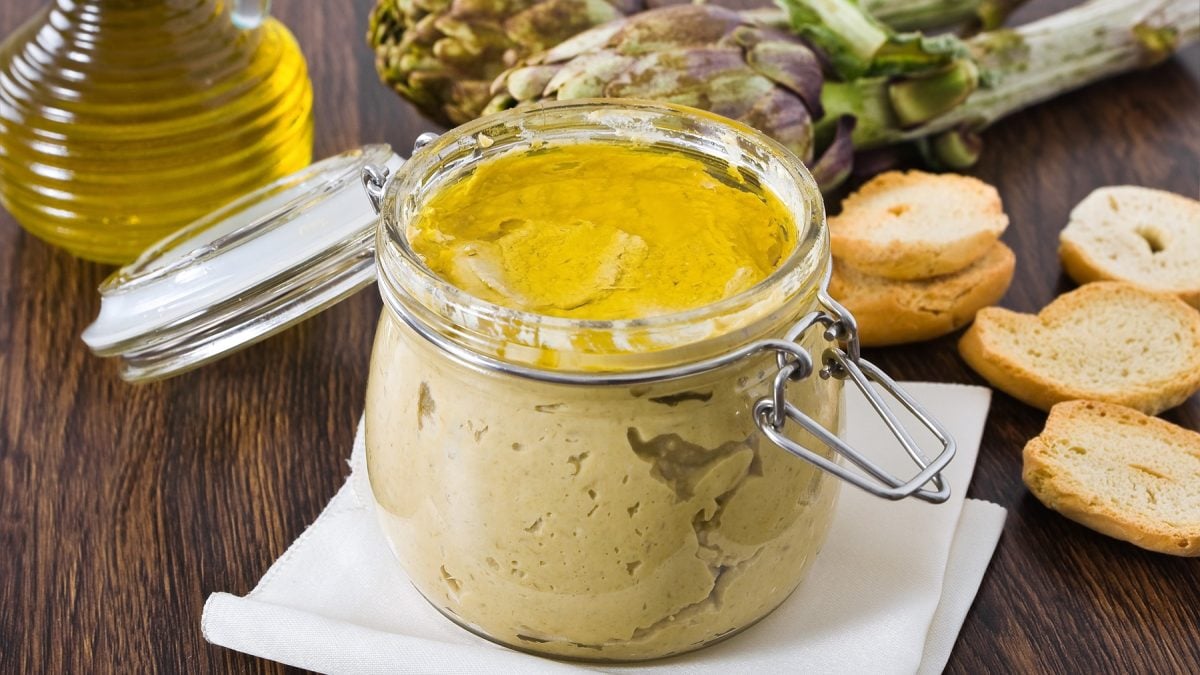 Offenes Glas mit cremigem Artischocken-Pesto, bedeckt mit Olivenöl, daneben geröstete Brotscheiben und frische Artischocken.