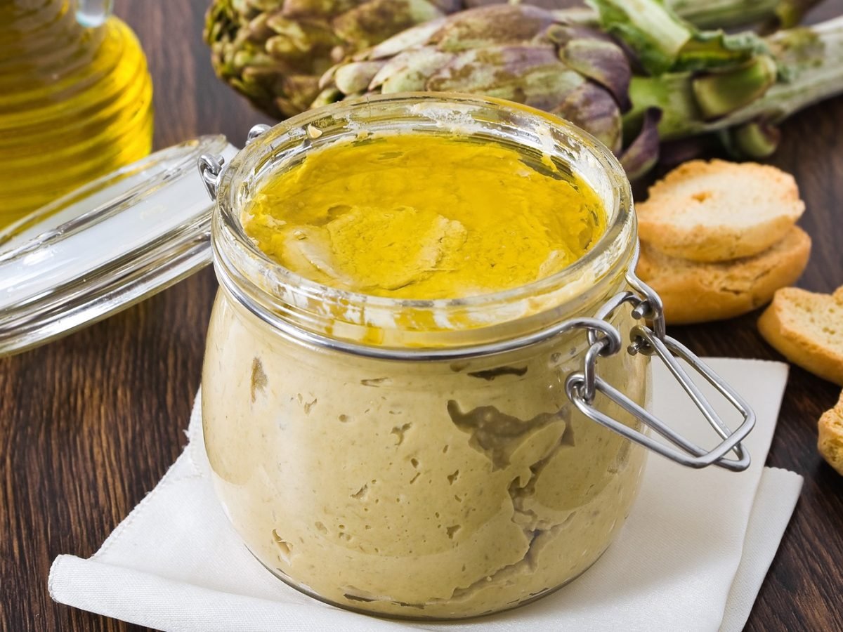 Offenes Glas mit cremigem Artischocken-Pesto, bedeckt mit Olivenöl, daneben geröstete Brotscheiben und frische Artischocken.