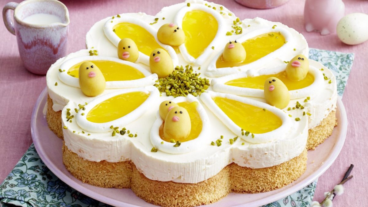 Draufsicht, schräg: Ein flache Torte auf einem runden Teller. Drum herum liegen zwei frische Eier. Daneben steht auf ein kleines Gefäß mit einem Henkel in dem eine helle Flüssigkeit ist.