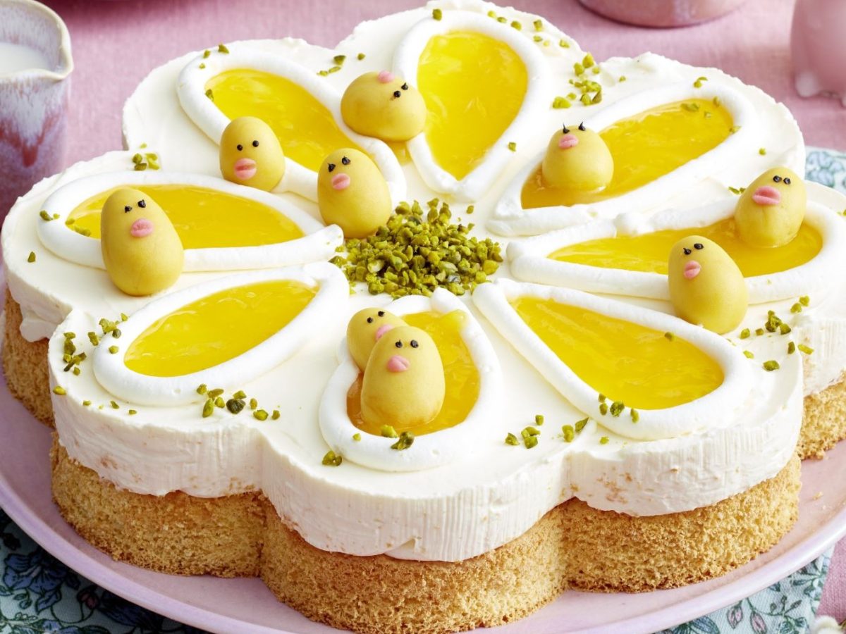 Draufsicht, schräg: Ein flache Torte auf einem runden Teller. Drum herum liegen zwei frische Eier. Daneben steht auf ein kleines Gefäß mit einem Henkel in dem eine helle Flüssigkeit ist.