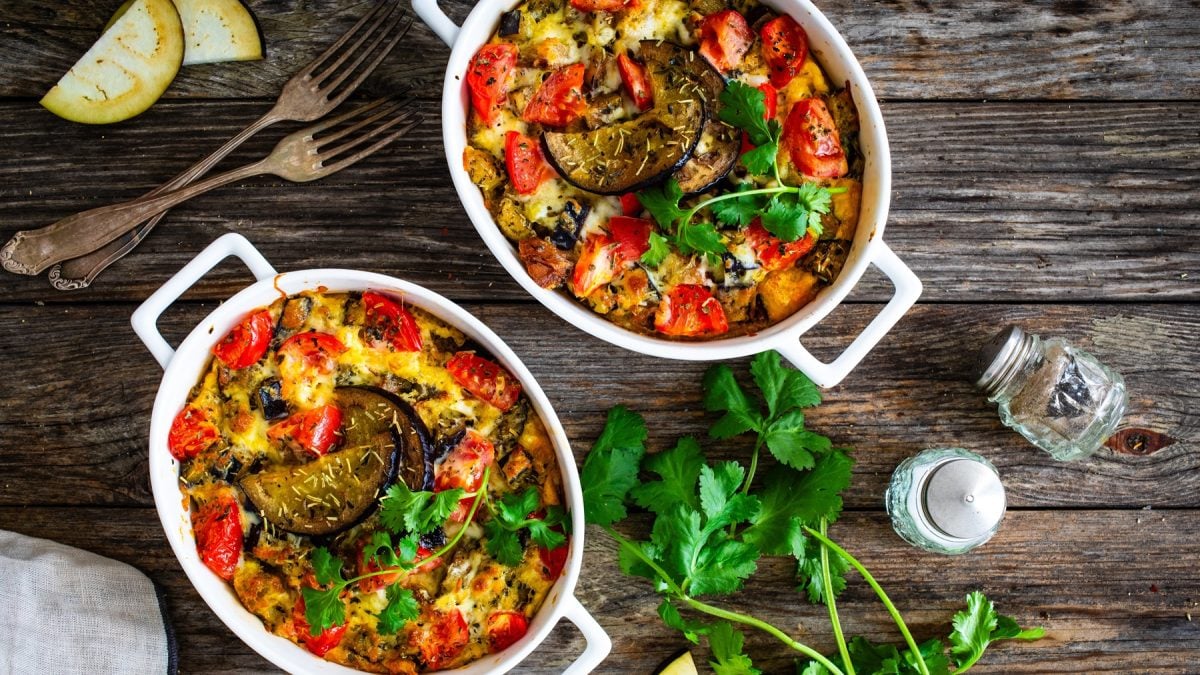 Zwei weiße Auflaufformen mit überbackenem Gemüse, frischen Tomaten, Auberginen und Kräutern auf rustikalem Holztisch.