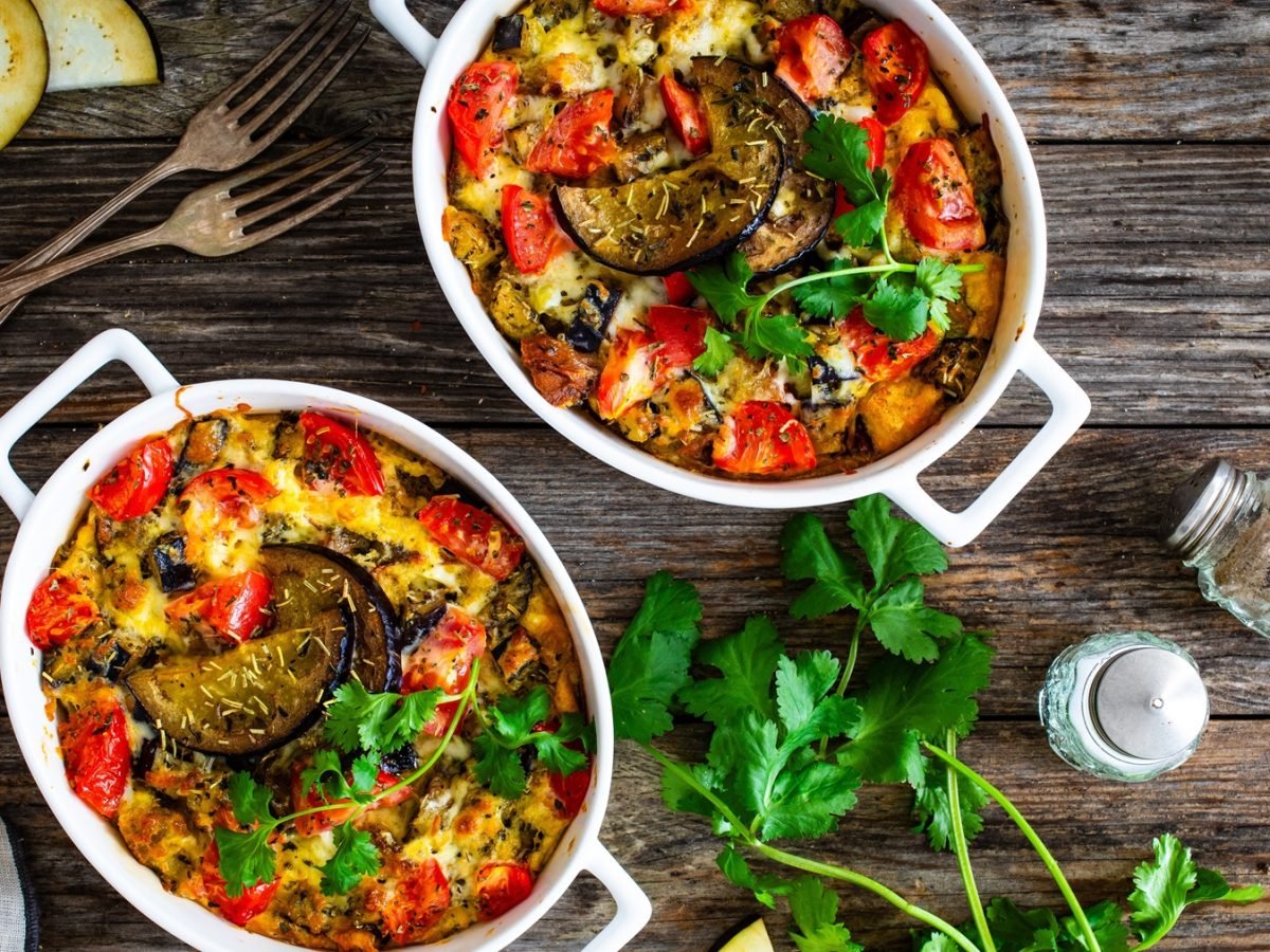 Zwei weiße Auflaufformen mit überbackenem Gemüse, frischen Tomaten, Auberginen und Kräutern auf rustikalem Holztisch.