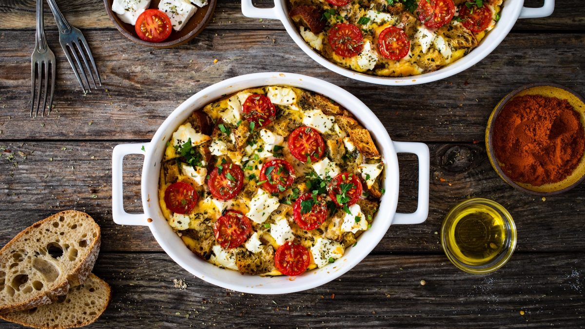 Überbackener Brotauflauf mit Tomaten, Feta und Kräutern in weißer Auflaufform auf rustikalem Holztisch serviert.