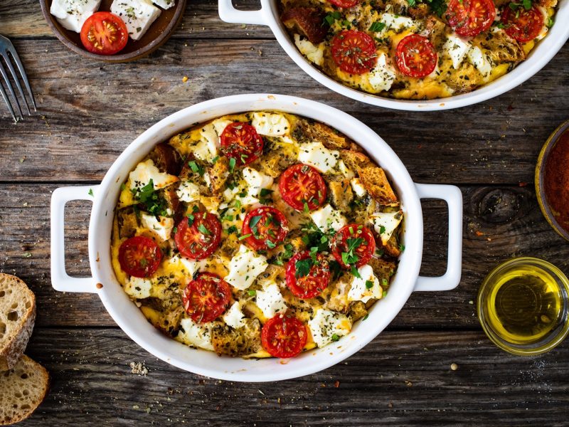 Überbackener Brotauflauf mit Tomaten, Feta und Kräutern in weißer Auflaufform auf rustikalem Holztisch serviert.