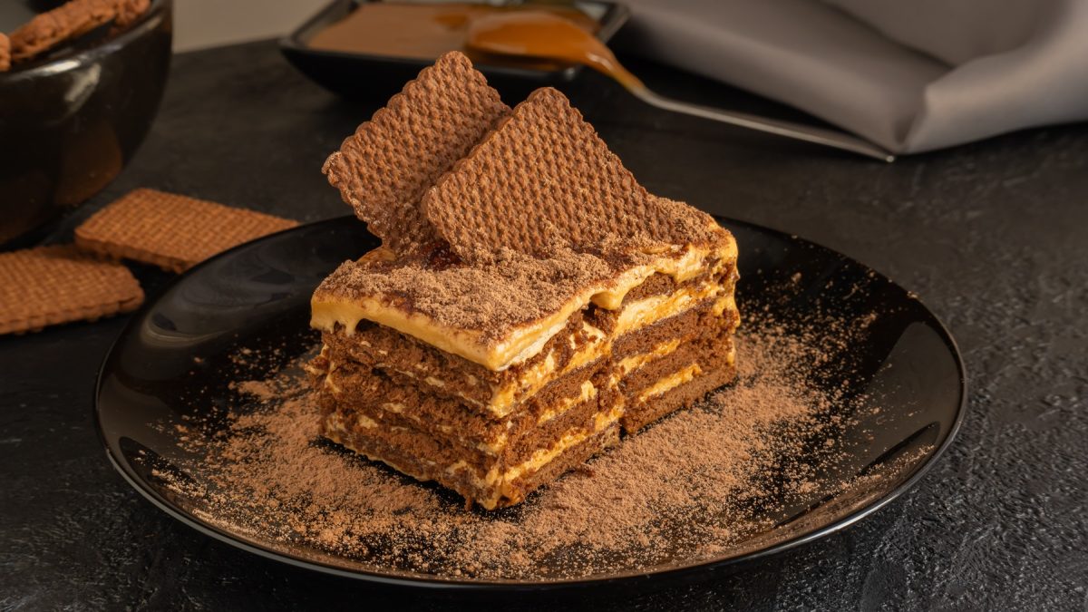 Nahaufnahme einer Chocotorta mit Kakaopulver und einer cremigen Füllung, dekoriert mit Waffeln, auf einem schwarzen Teller.