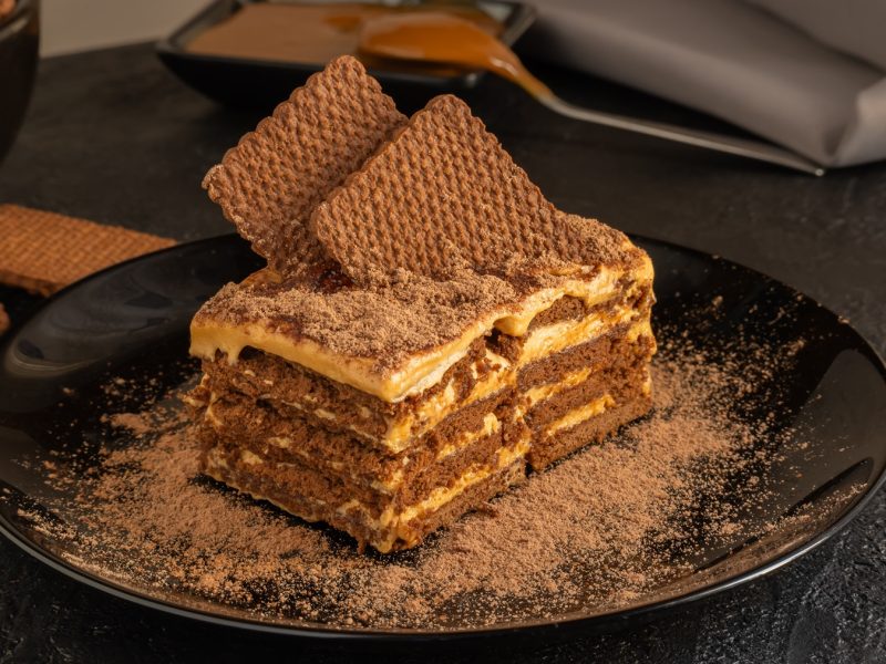 Nahaufnahme einer Chocotorta mit Kakaopulver und einer cremigen Füllung, dekoriert mit Waffeln, auf einem schwarzen Teller.