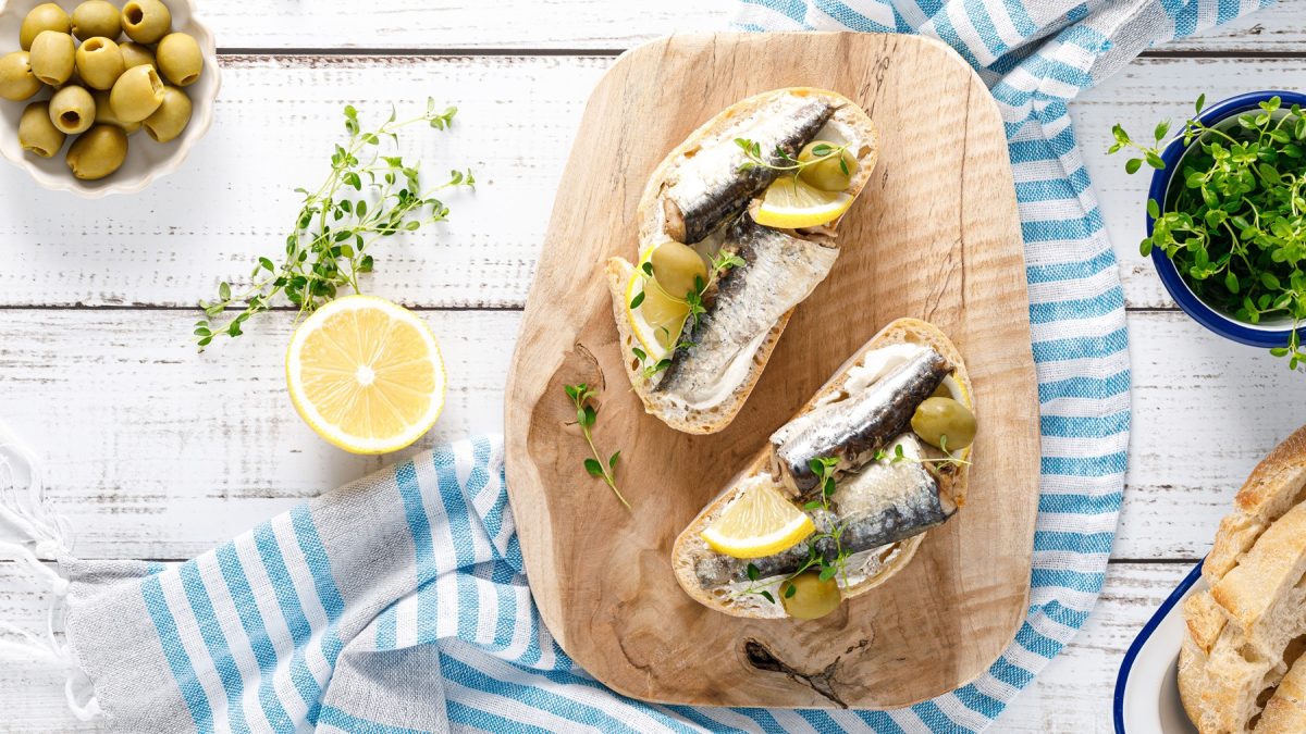 Zwei belegte Crostini mit Sardinen, Frischkäse, Oliven und Zitrone auf einem Holzbrett, umgeben von Kräutern auf rustikalem Holztisch.
