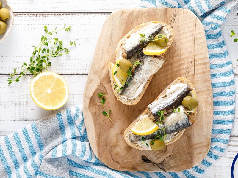 Zwei belegte Crostini mit Sardinen, Frischkäse, Oliven und Zitrone auf einem Holzbrett, umgeben von Kräutern auf rustikalem Holztisch.