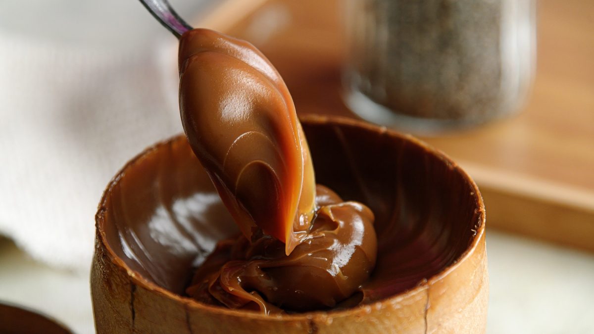 Nahaufnahme eines Löffels mit glänzendem Dulce de leche über einer hölzernen Schale vor unscharfem Hintergrund.