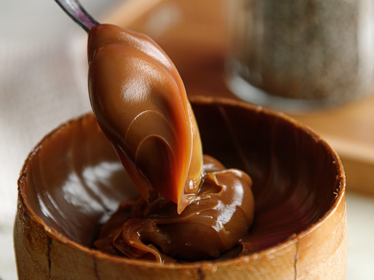 Nahaufnahme eines Löffels mit glänzendem Dulce de leche über einer hölzernen Schale vor unscharfem Hintergrund.