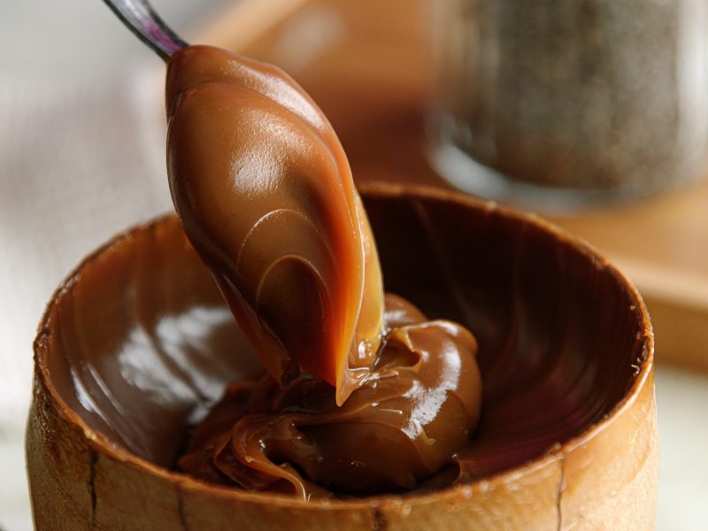 Nahaufnahme eines Löffels mit glänzendem Dulce de leche über einer hölzernen Schale vor unscharfem Hintergrund.